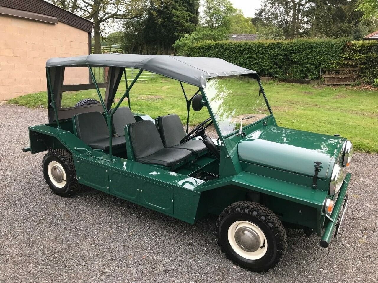 A green, open-top utility vehicle with four seats, rugged tires, and a simple frame is parked on gravel near grass and trees.