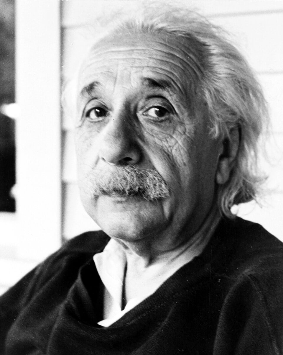 An older man with white hair and a mustache looks at the camera, wearing a dark sweater over a collared shirt, sitting in front of a light-colored wall.