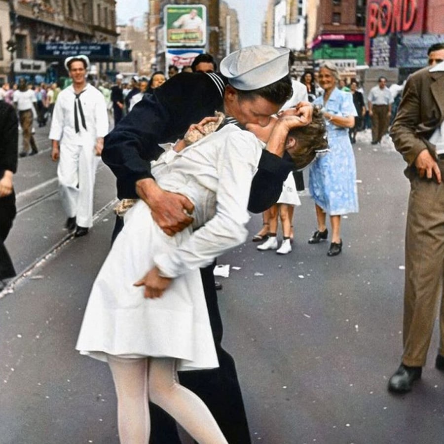 A sailor kisses a woman in a white dress on a busy city street, surrounded by people celebrating. The scene appears joyful and lively, likely during a public celebration.