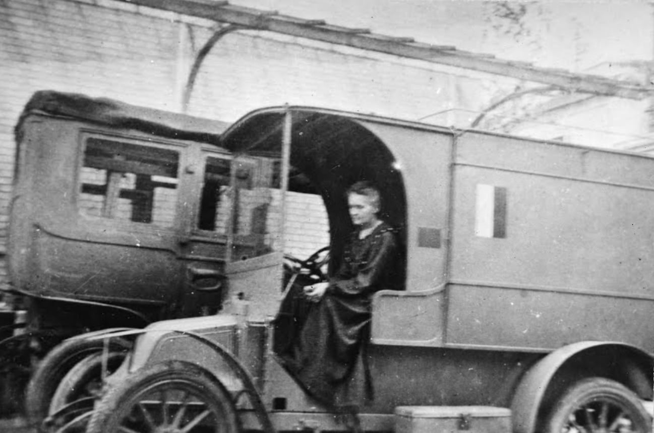 A woman in a long coat sits in the driver’s seat of an old-fashioned ambulance van, with another vehicle parked beside it, both in front of a brick building.