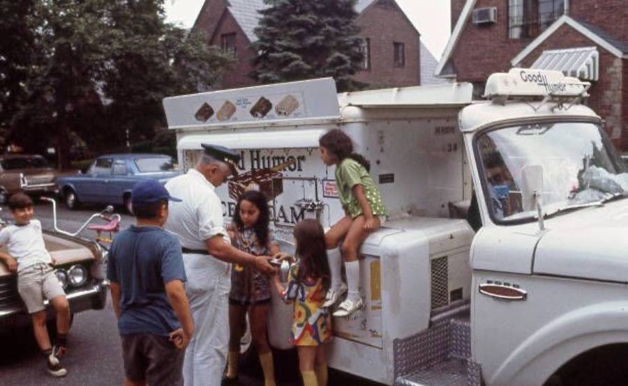 Children buy ice cream from a man in uniform at a vintage ice cream truck parked on a suburban street. Some kids sit on the truck while others stand nearby, enjoying the summer day.