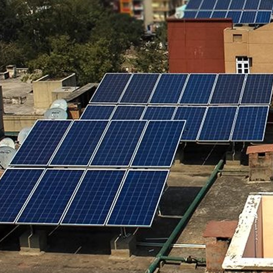 Rows of blue solar panels are installed on a flat rooftop in an urban area, with nearby buildings and trees in the background.
