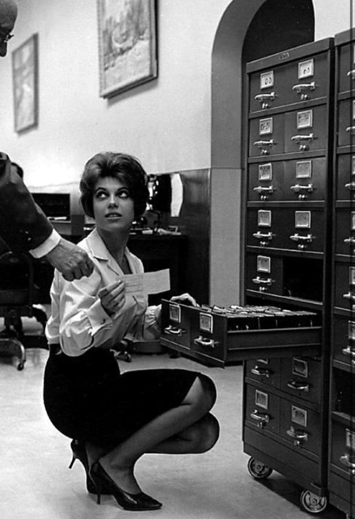 A woman in a skirt and heels kneels by an open file cabinet drawer, holding an index card and looking up at a person’s outstretched hand in an office setting.