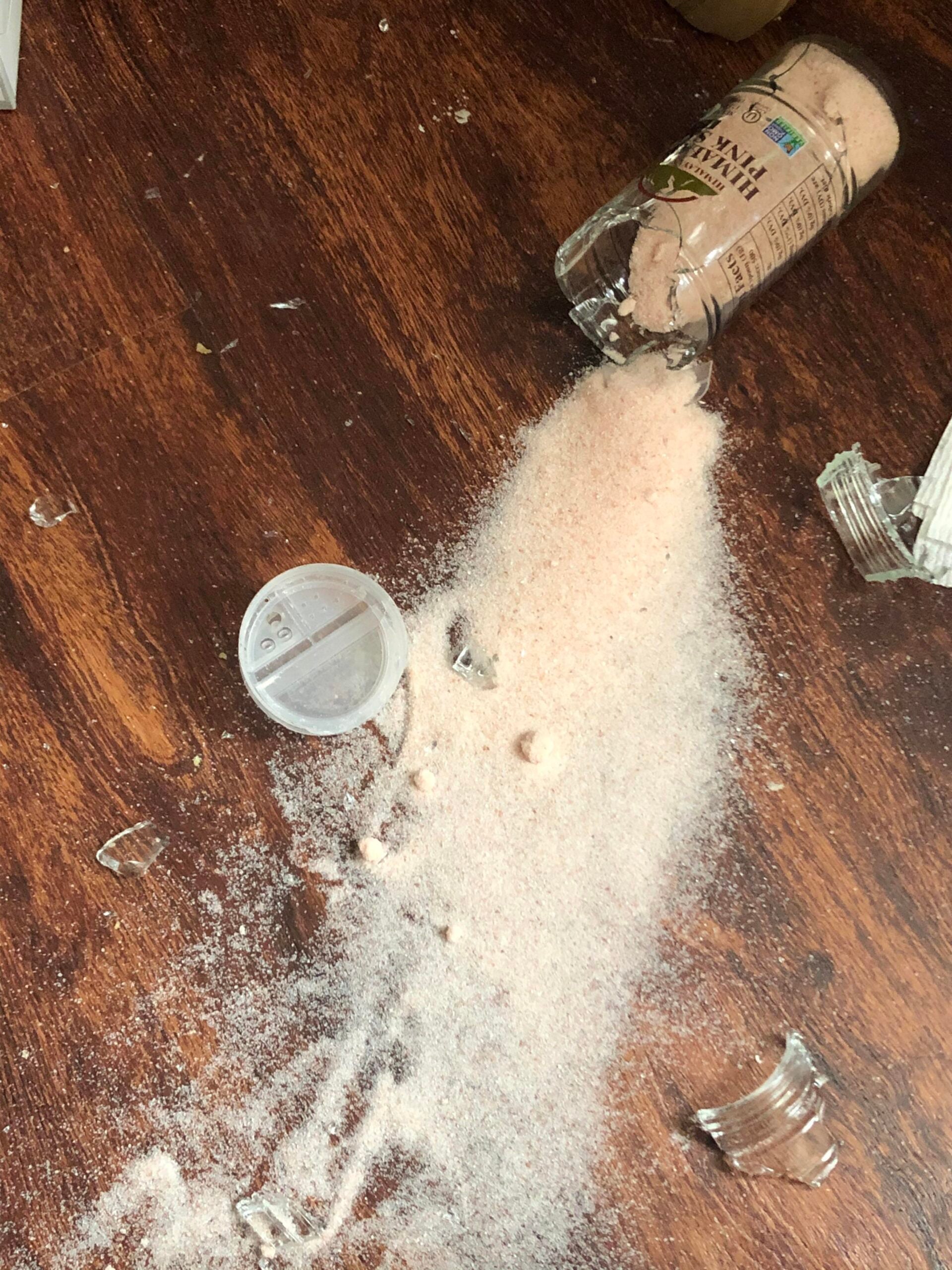 A glass container of pink Himalayan salt has broken and spilled salt and glass shards across a wooden floor. The salt cap is off and lying nearby.