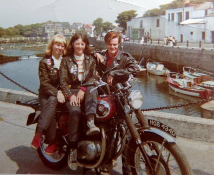 Three people in leather jackets sit on a motorcycle by a waterfront, with boats and buildings in the background. The person in front is holding the handlebars, while the other two smile at the camera.