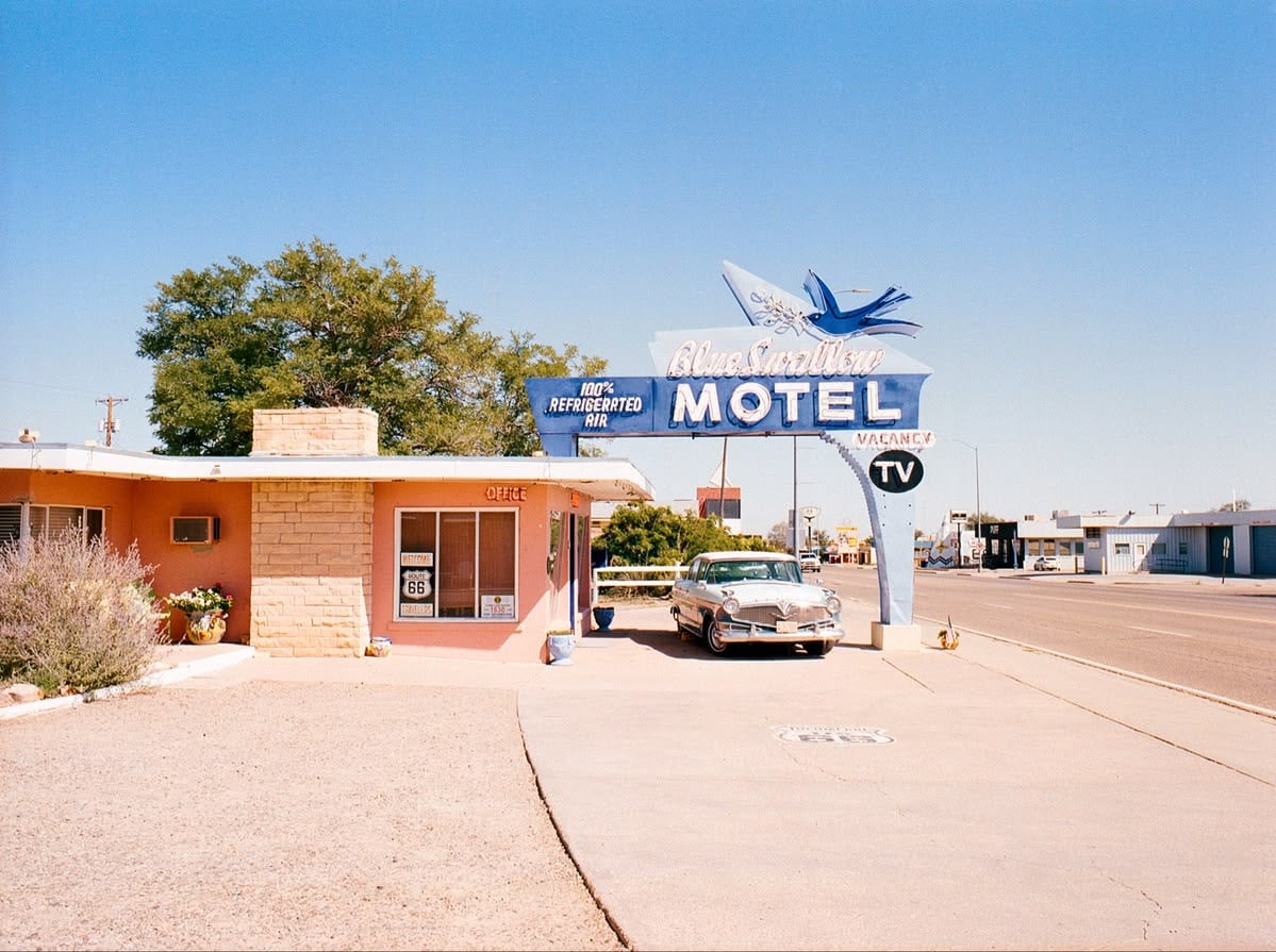 A vintage car is parked outside the Blue Swallow Motel, featuring a neon sign and retro architecture, under a clear blue sky on a quiet street.