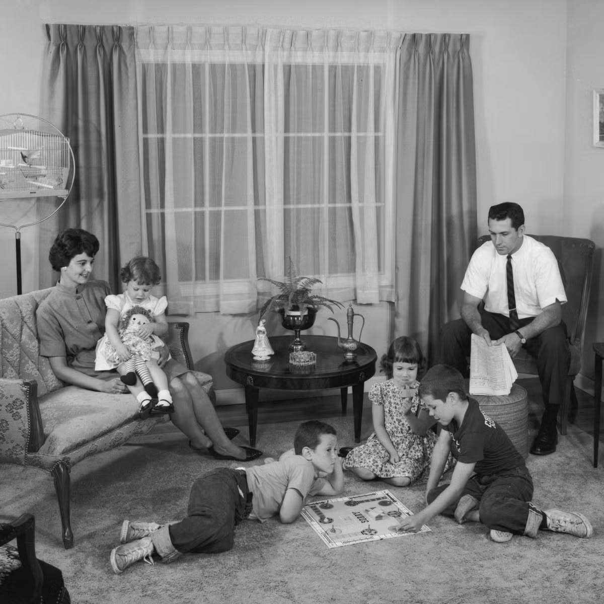 A family sits in a living room: a woman with a doll-holding girl, a man with a newspaper, and three kids playing a board game on the floor. The room is decorated with curtains, a table, and a birdcage.