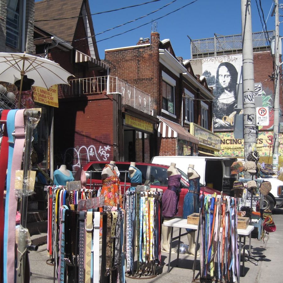 Outdoor market scene with tables of colorful belts and scarves. Mannequins display accessories. Brick buildings and shops are in the background, with street art and a mural resembling the Mona Lisa on the wall.