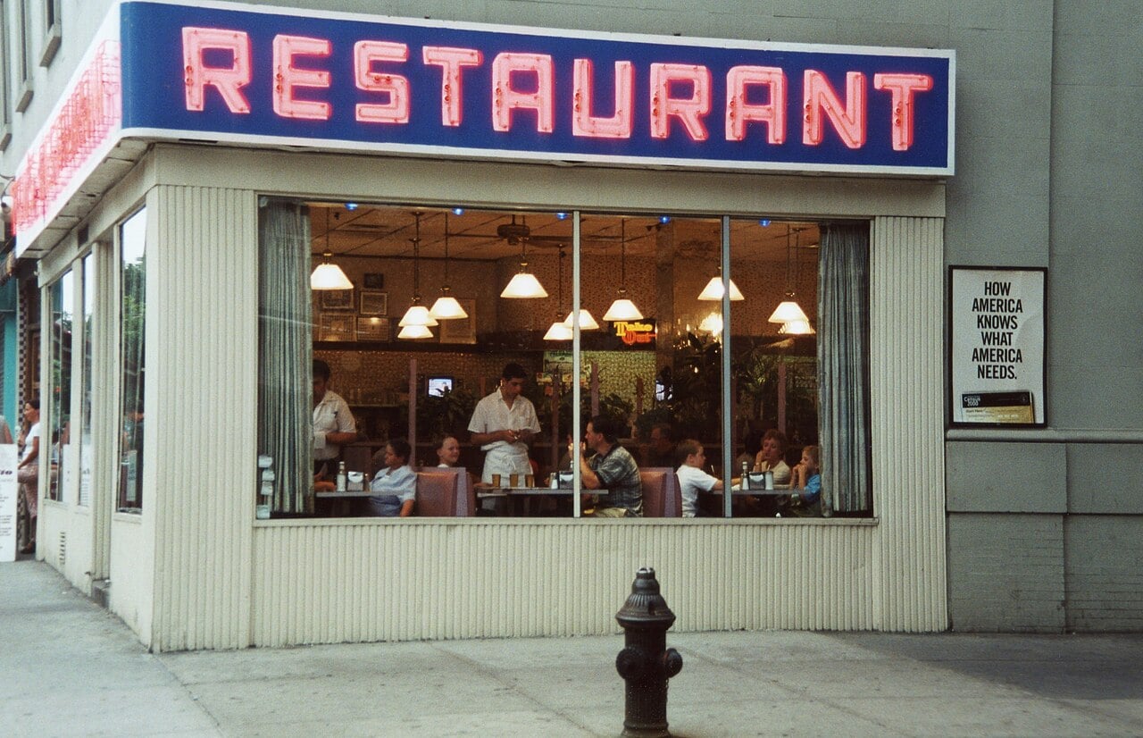 Street view of a diner with a neon “RESTAURANT” sign. People sit inside at tables, and a server is taking orders. A sign on the wall reads, “HOW AMERICA KNOWS WHAT AMERICA NEEDS.” A fire hydrant is visible on the sidewalk.