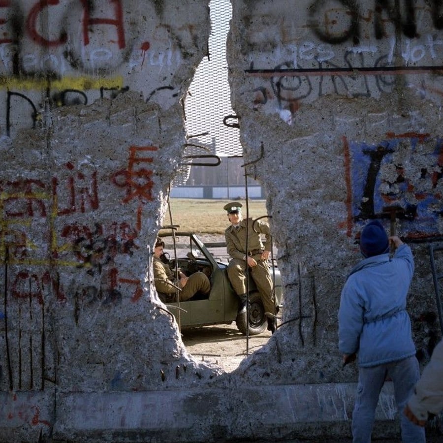 A person in a blue jacket looks through a large hole in a graffiti-covered concrete wall as two uniformed guards sit in a military vehicle on the other side.