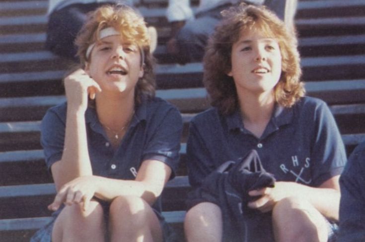 Two teens with short hair sit on outdoor bleachers, wearing matching blue polo shirts with “RHS” and tennis racquet logos. One has a headband and rests her chin on her hand, both are smiling.