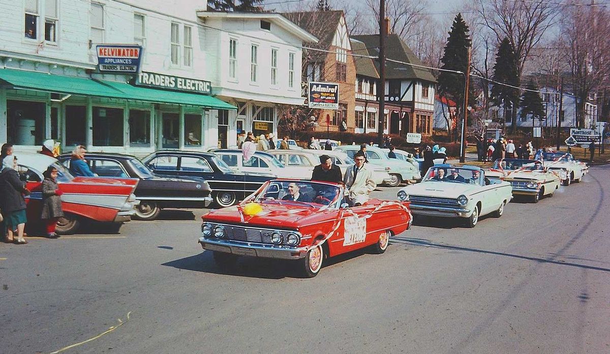 A colorful parade of vintage cars drives down a street lined with spectators, shops, and houses. People watch from the sidewalk as participants ride in the decorated convertibles.