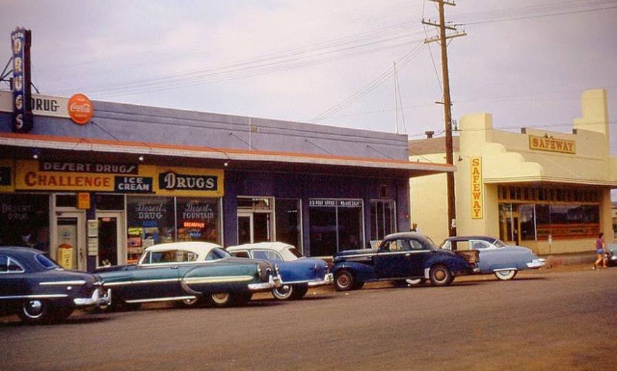 A vintage street scene with classic cars parked in front of a drugstore and a Safeway grocery store. The storefronts have retro signs, and the overall atmosphere reflects a mid-20th century small town.