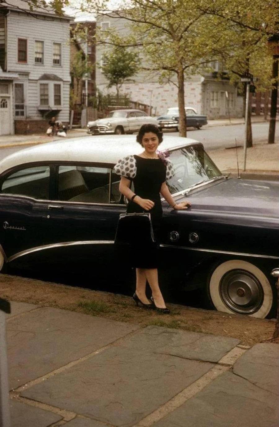 A woman in a black dress with polka-dot sleeves stands on a sidewalk next to a vintage black car, holding a handbag. Trees, houses, and a quiet street are visible in the background.