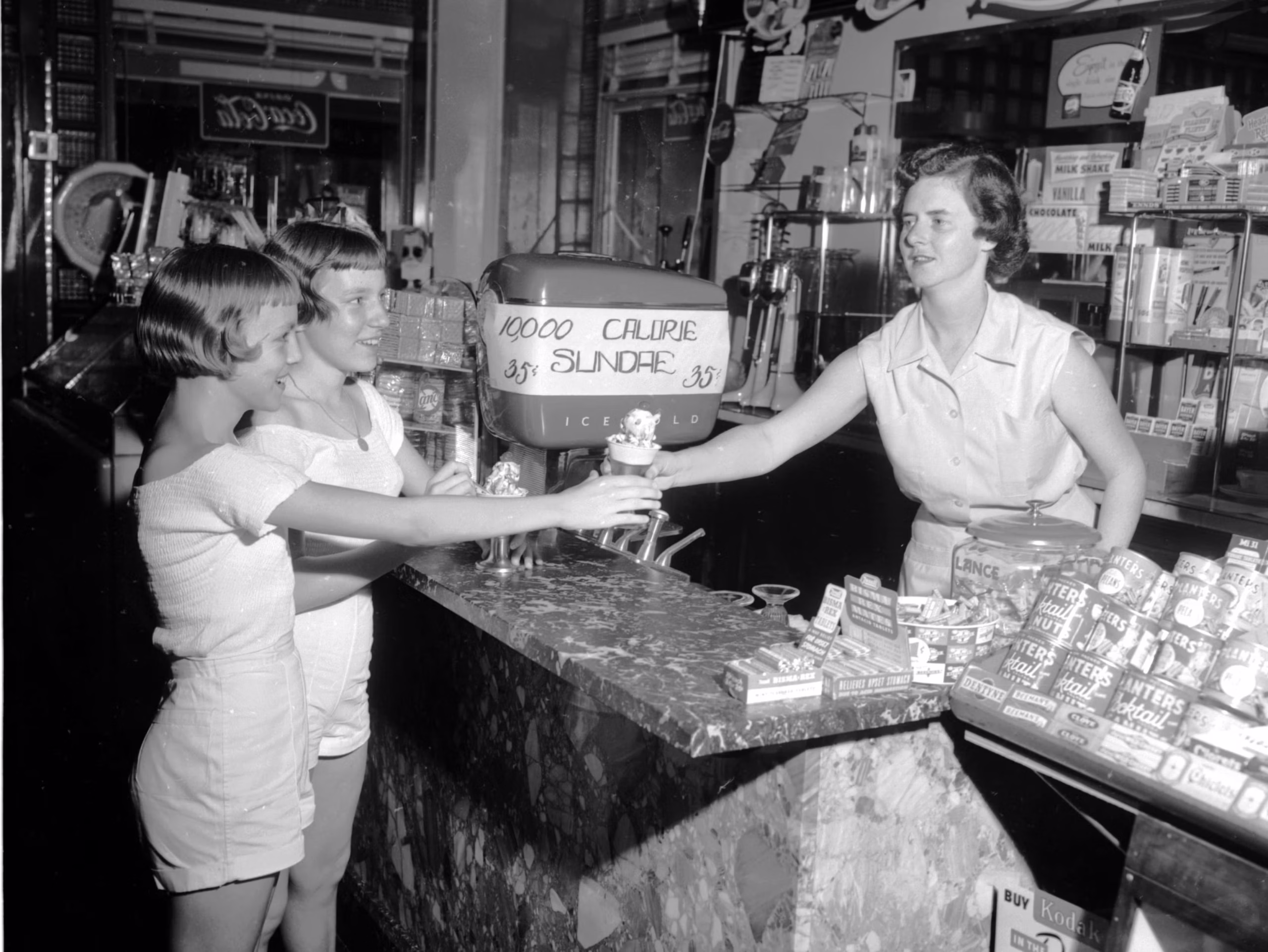 A woman behind a counter hands ice cream sundaes to two smiling girls. A sign advertises a “10,000 Calorie Sundae.” The counter is filled with various candy and snack products. The scene appears to be from the 1950s.