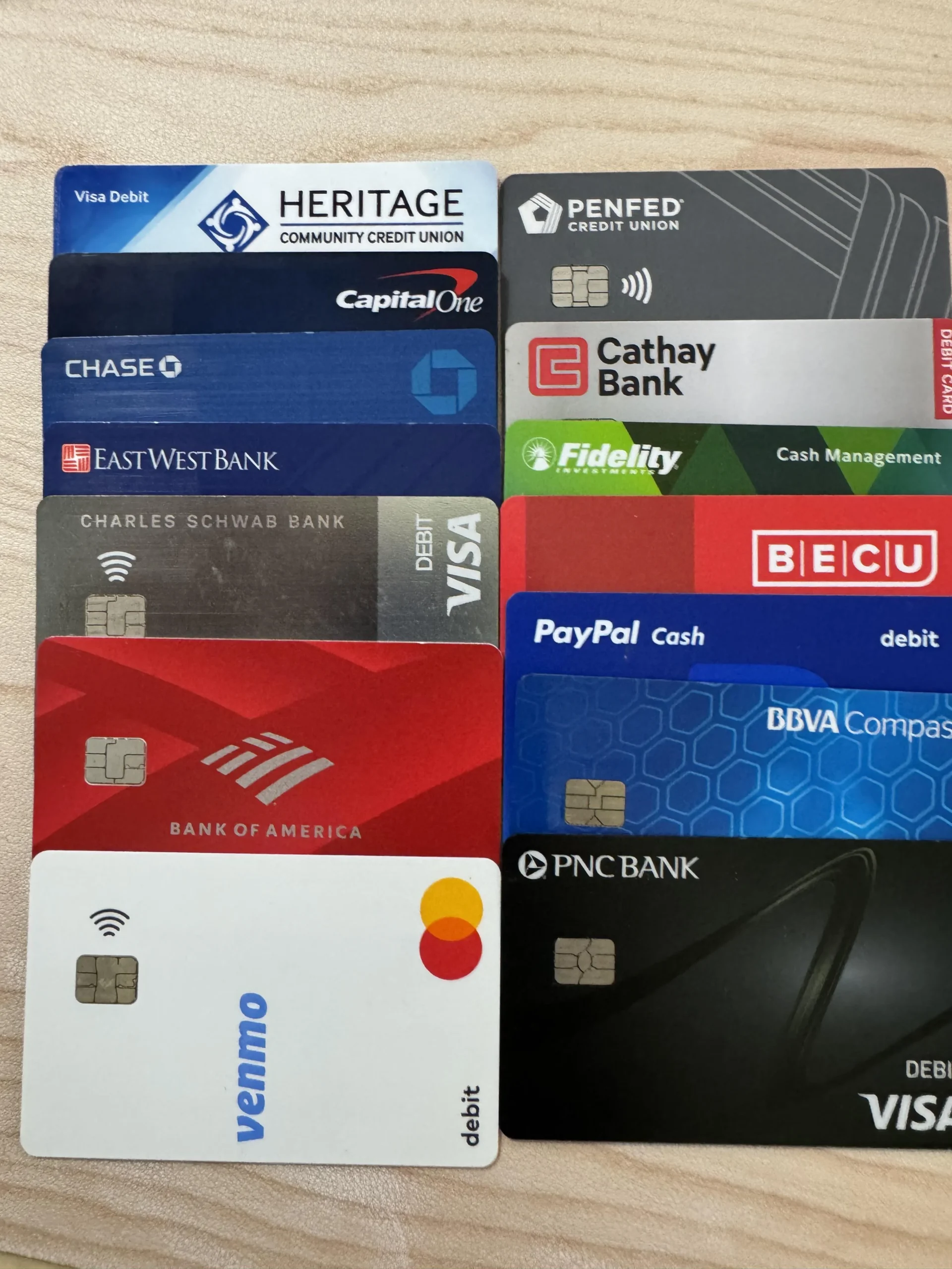 A collection of twelve assorted debit and credit cards from various banks and financial institutions arranged neatly on a light wood surface.