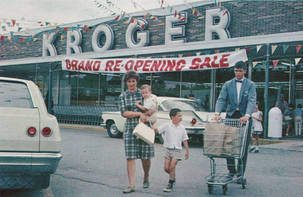 A family leaves a Kroger supermarket during a grand re-opening sale. The mother carries a baby and holds another child's hand, while a man pushes a cart with groceries in the parking lot. 1960s cars are parked outside.
