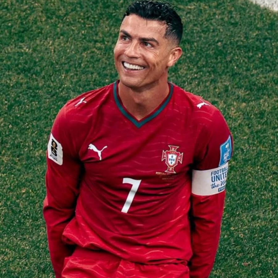 A soccer player in a red Portugal jersey with the number 7 smiles while standing on a grassy field. He has short dark hair and is wearing a captain's armband.