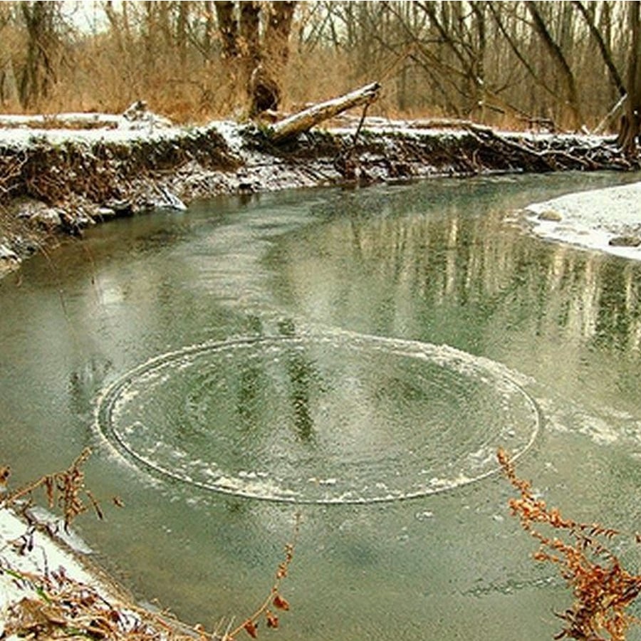 A partially frozen river with a perfect circular formation of ice near the shore, surrounded by snow-dusted trees and fallen branches.