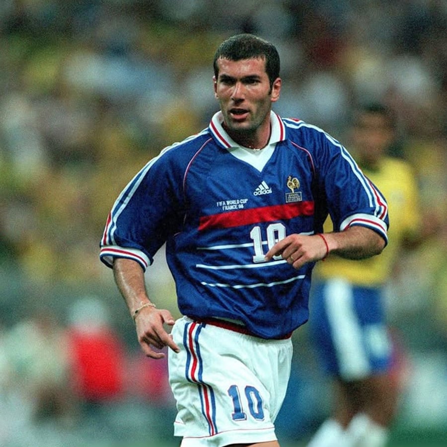 A soccer player in a blue France jersey with number 10 runs on the field during a match. The background is blurred, showing other players and a crowd.