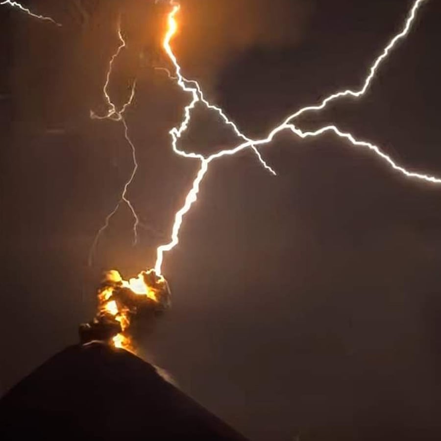 A bright lightning bolt strikes the erupting peak of a volcano at night, illuminating clouds of ash and lava against a dark sky.