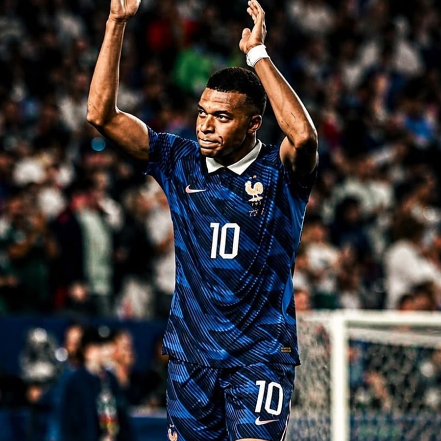 A soccer player in a blue France jersey with the number 10 claps above his head on the field, with a crowd blurred in the background.