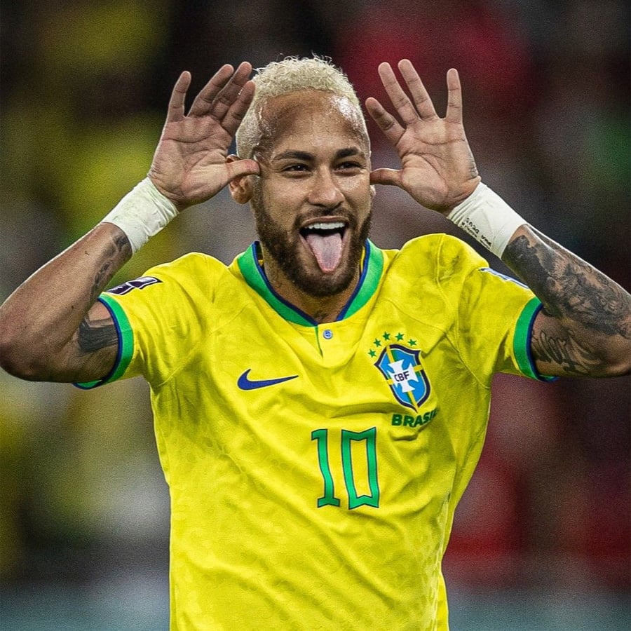 A soccer player in a yellow Brazil jersey with number 10 makes a playful face, sticking out his tongue and holding his hands up by his head with splayed fingers.