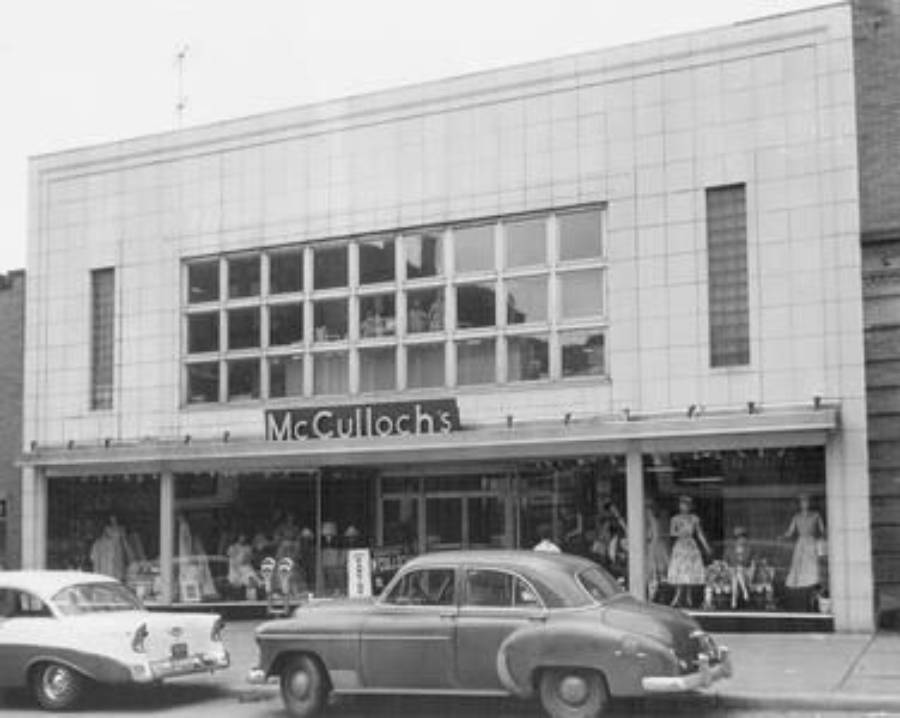Black and white photo of McCulloch’s storefront with large display windows; vintage cars are parked in front and mannequins wearing dresses are visible in the window.