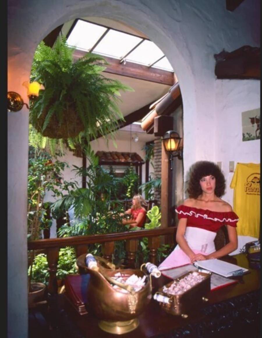 A woman in a red and white off-shoulder dress stands behind a desk with champagne and a guestbook, in a lush, plant-filled room with arched walls and natural light.