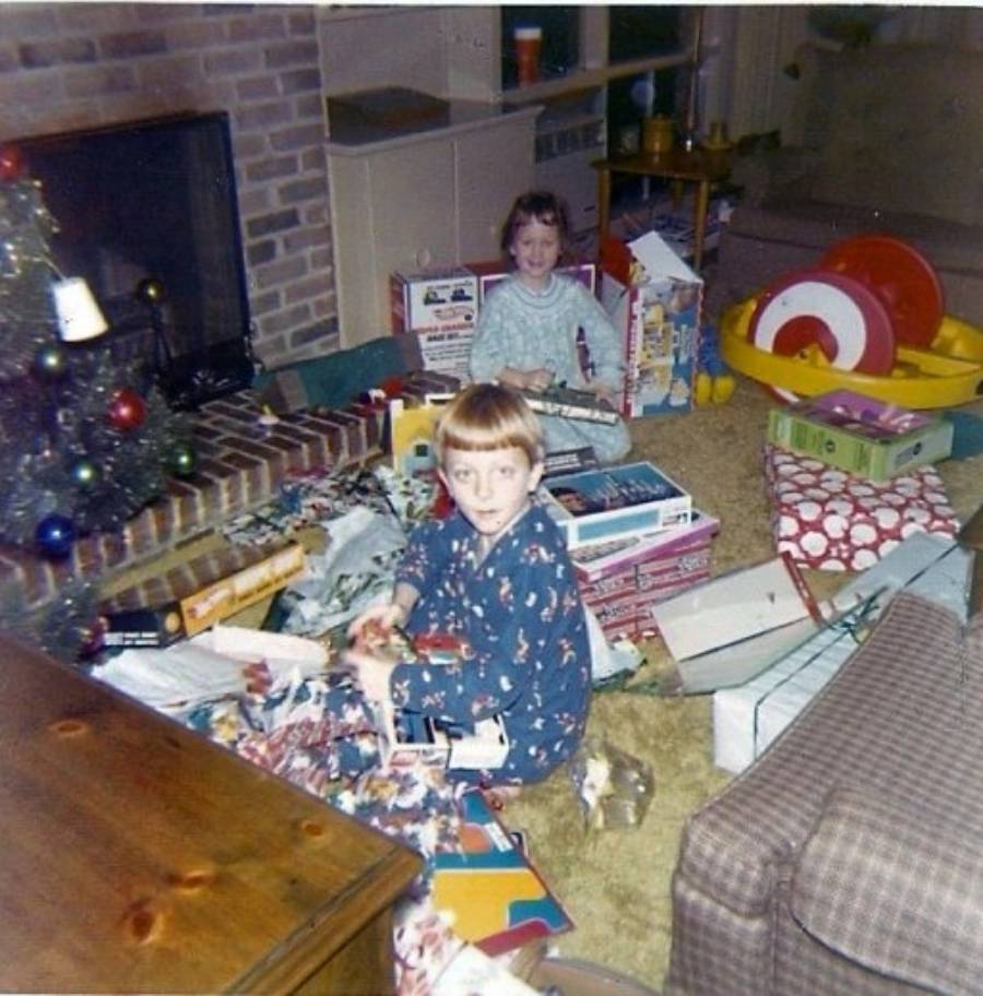 Two children in pajamas sit among opened presents and wrapping paper near a Christmas tree in a cozy living room, smiling at the camera. A fireplace and various toys are visible in the background.