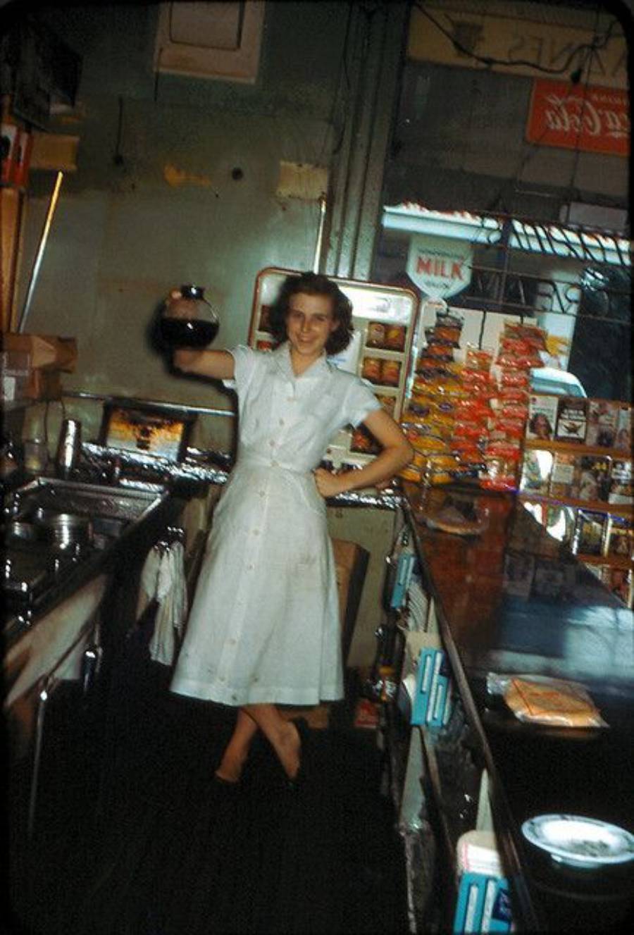 A woman in a white dress stands behind a diner counter, smiling and holding a coffee pot. The background includes a milk machine, snacks, and various diner items.