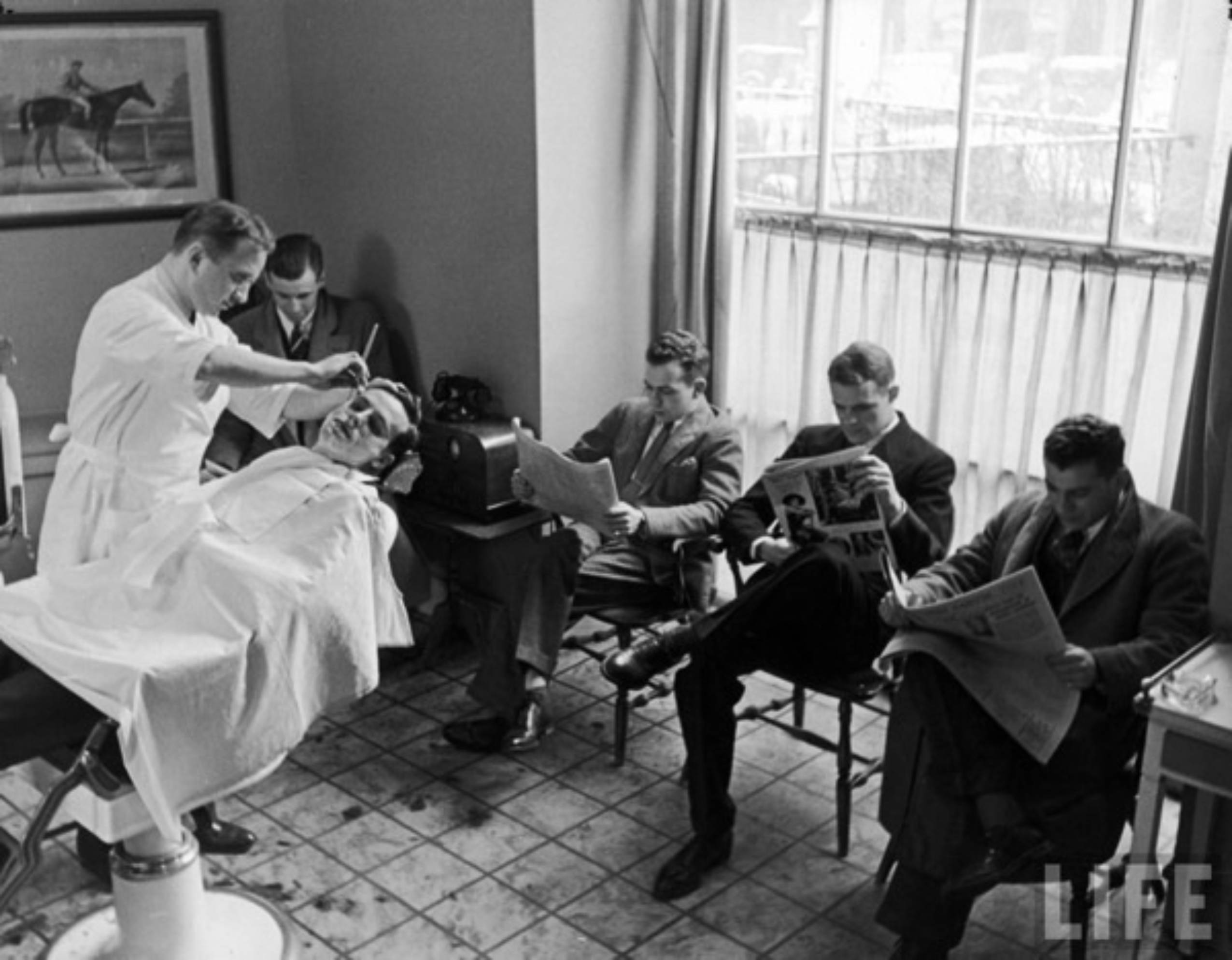 A barber shaves a man in a chair while four men wait, reading newspapers and magazines in a vintage barbershop with tiled floors and a large window.