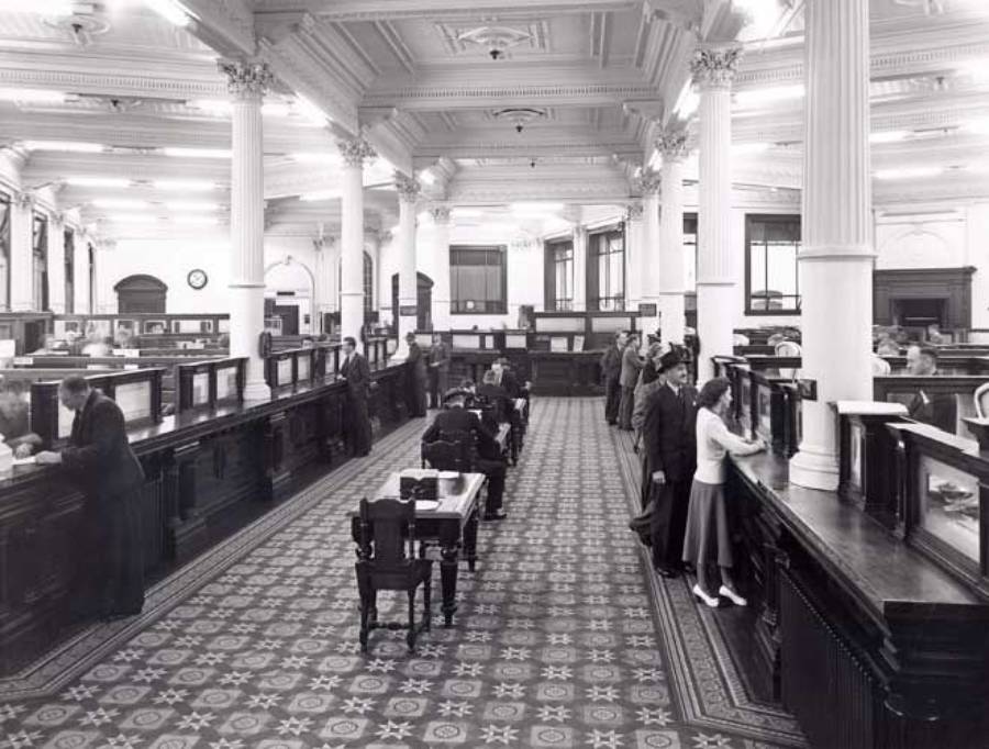 Black and white photo of a spacious, ornate bank interior with people standing at counters and sitting at desks under high ceilings and large columns. The patterned floor and classic architecture suggest an early 20th-century setting.