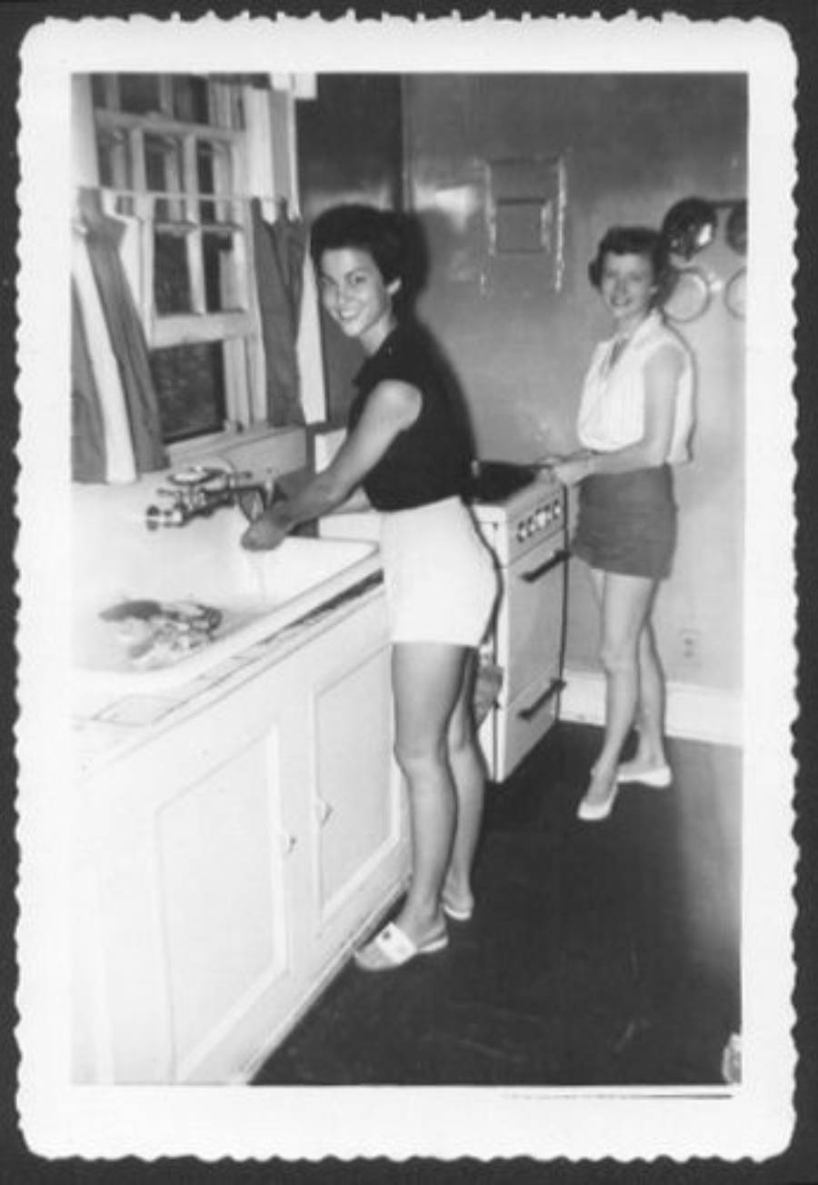 Two women in vintage clothing stand in a kitchen. One washes dishes at the sink, smiling at the camera, while the other stands by a stove in the background. The photo has scalloped edges and a black-and-white filter.