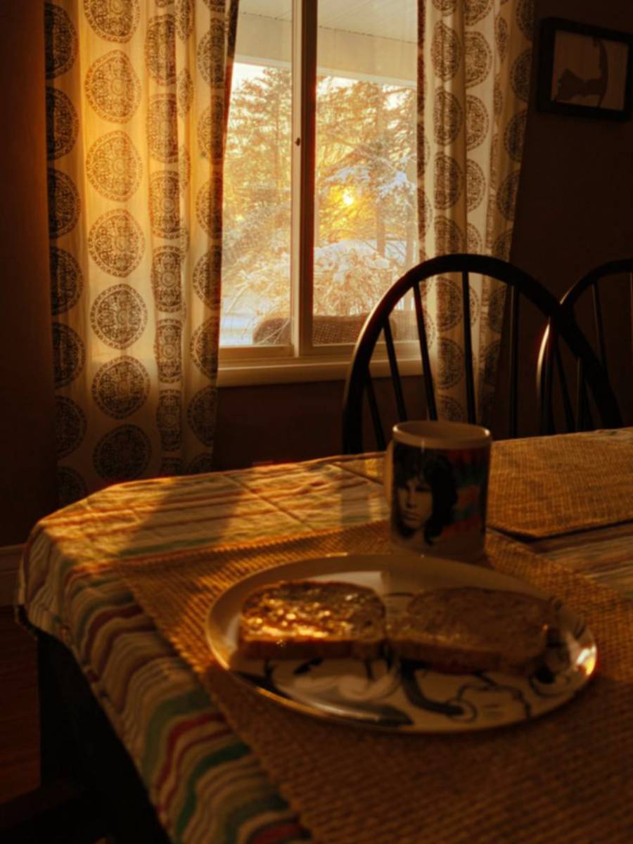 A breakfast table with two slices of toast on a plate and a mug. Warm sunlight streams through a window with curtains, casting golden light across the room. Snow and trees are visible outside the window.