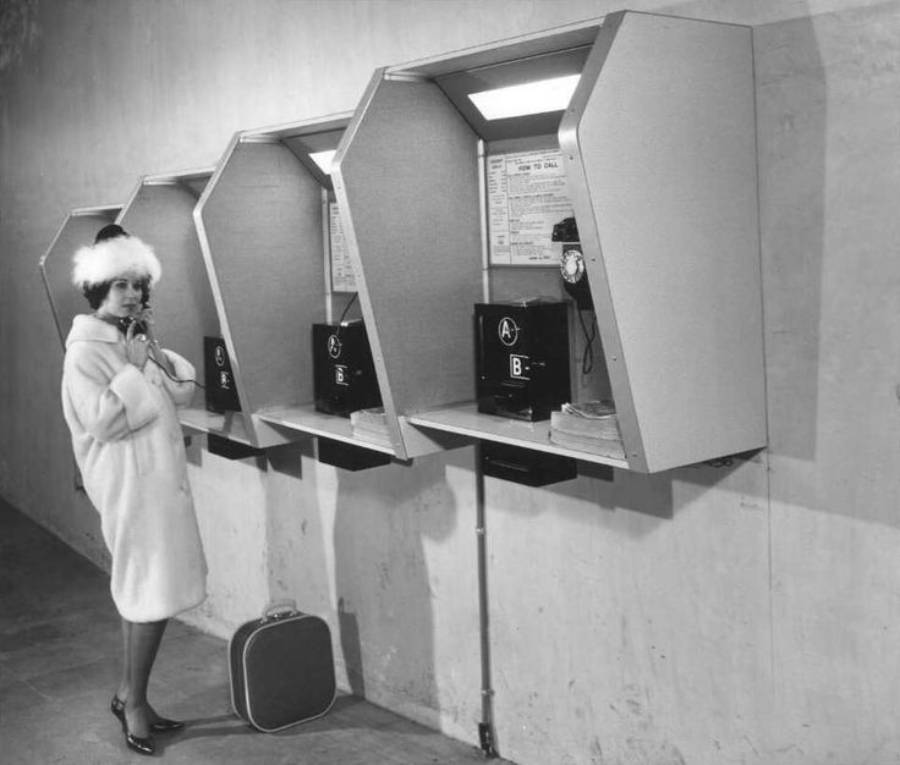 A woman in a white coat and hat stands by vintage payphones, holding a receiver to her ear. A small suitcase is on the floor next to her. The setting looks like an old airport or station.