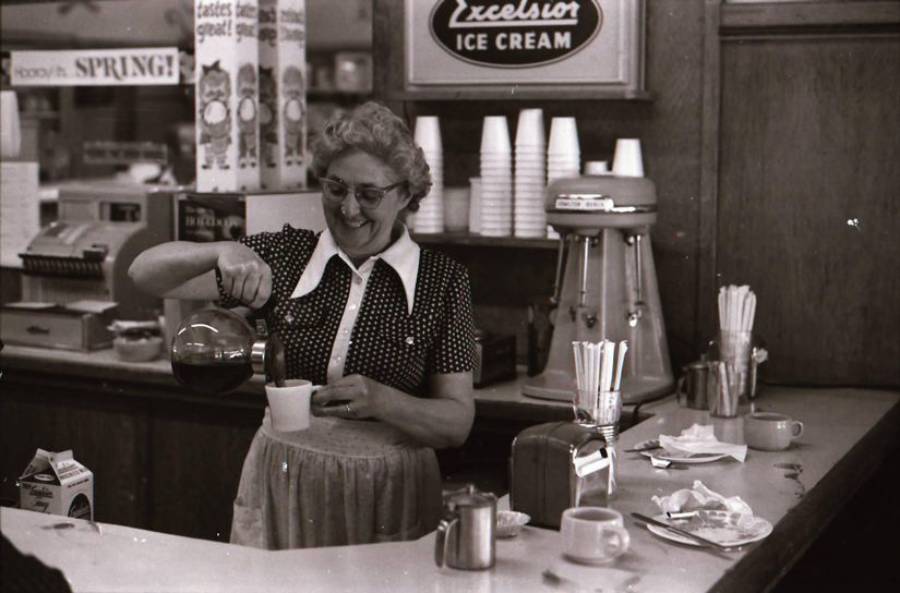 Smiling woman in a polka dot dress and apron pours coffee behind a vintage diner counter with cups, a milk carton, a napkin holder, and an “Excelsior Ice Cream” sign in the background.