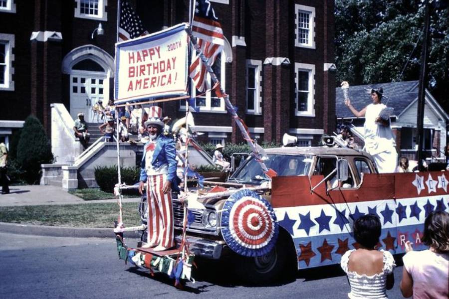 A parade float decorated with stars, stripes, and a “Happy Birthday America!” sign, featuring people dressed in patriotic attire, passes by spectators in front of a brick building.