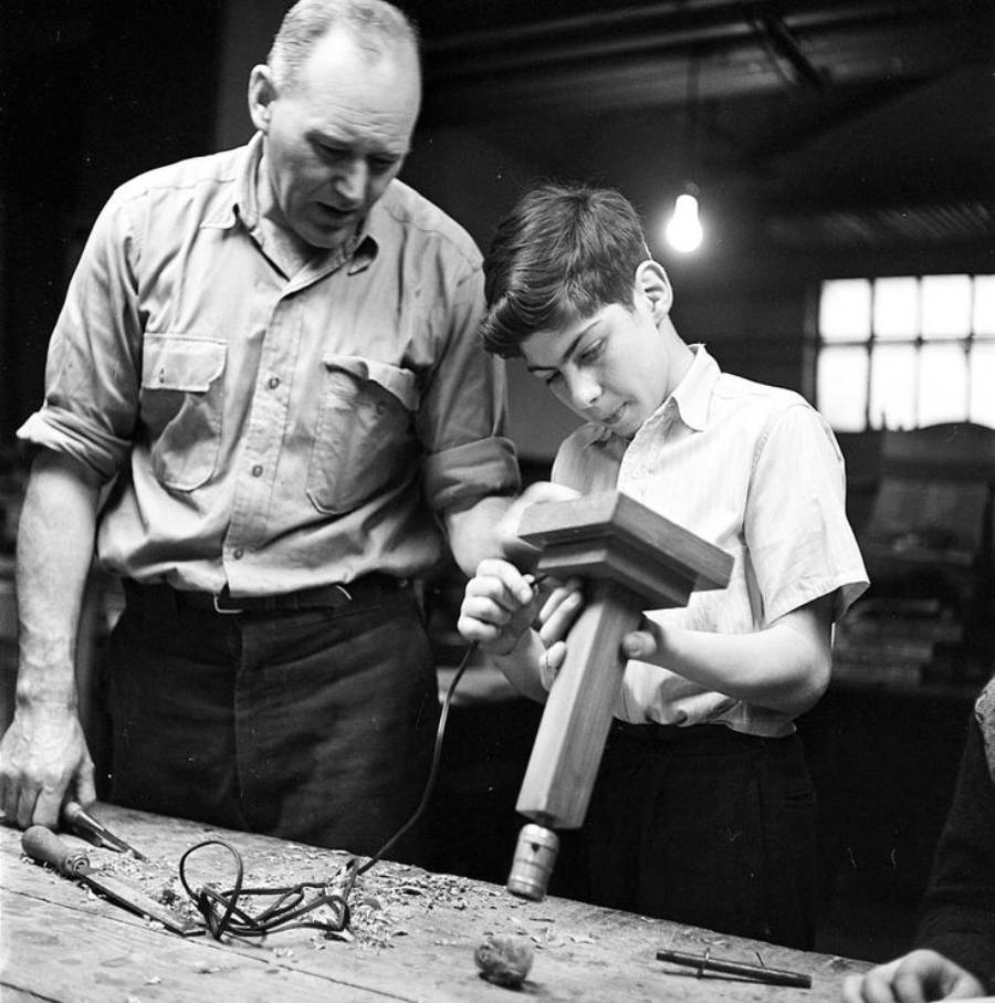 A man watches as a boy uses a power drill on a wooden object at a workbench, surrounded by tools, in a workshop with overhead lights.