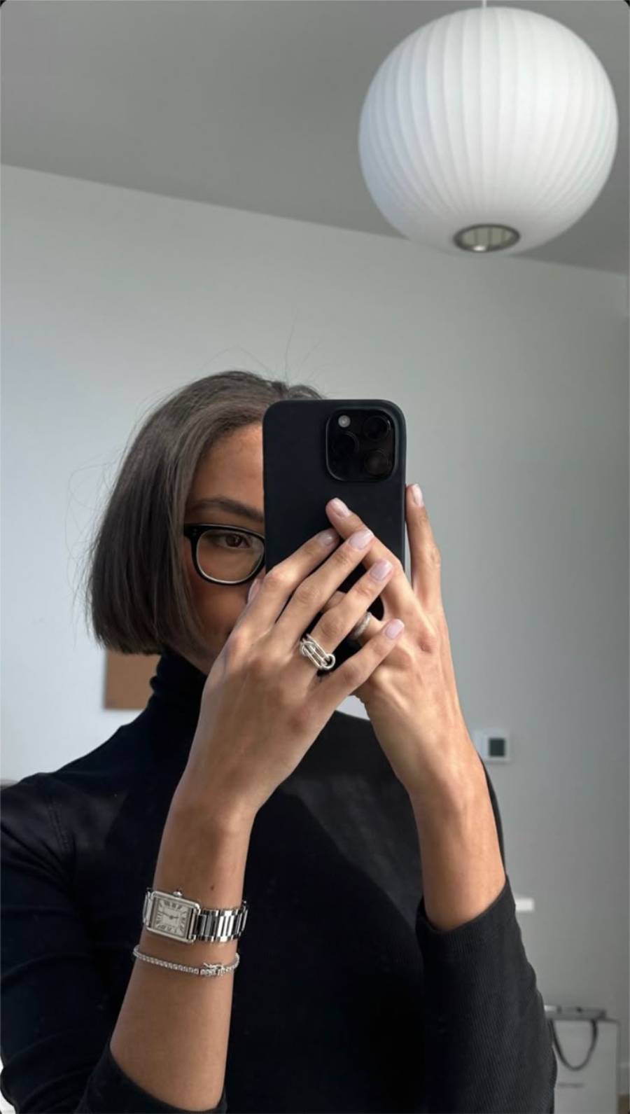 Person with a bob haircut and glasses takes a mirror selfie with a black phone, wearing a black turtleneck, silver watch, and ring. A round white ceiling lamp is visible above.