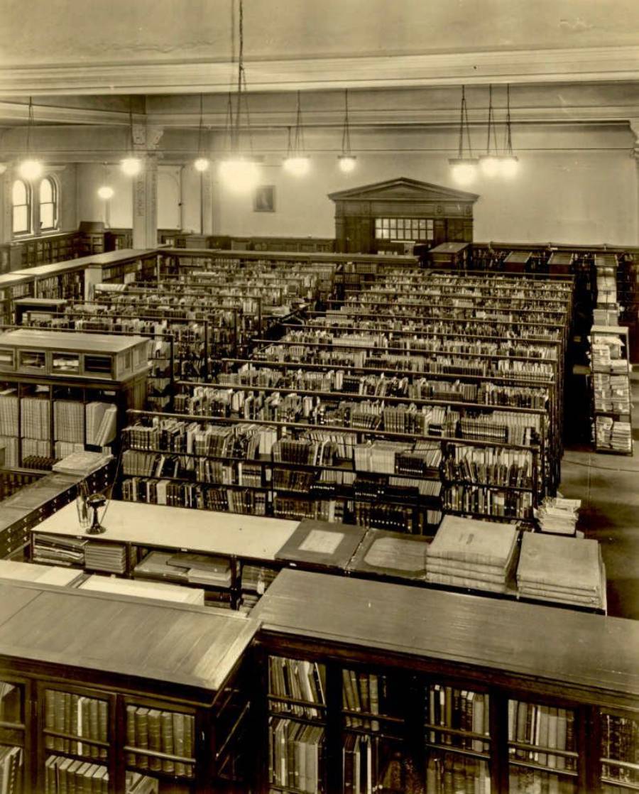 Historic black-and-white photo of a large library room filled with rows of bookshelves, tables with books, and tall windows letting in natural light. Overhead lights hang from the high ceiling.