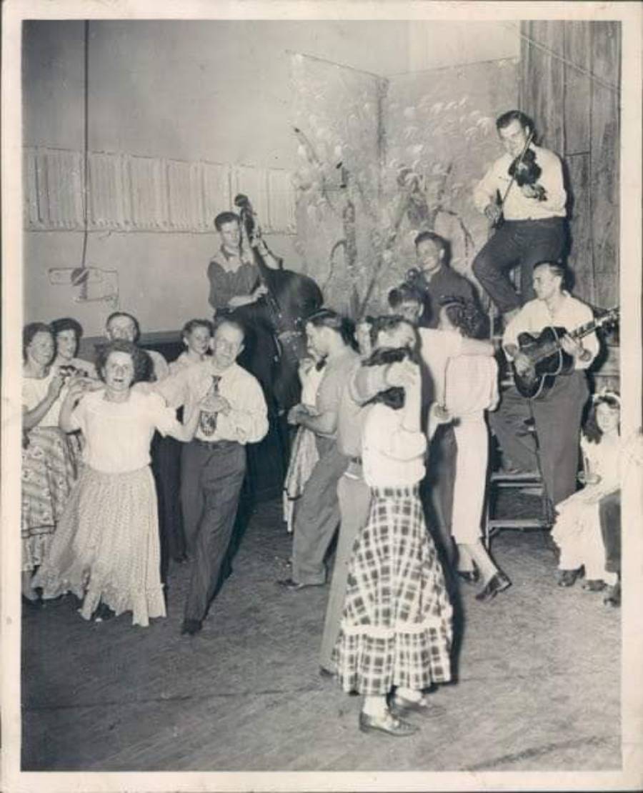 Black-and-white photo of people square dancing in a hall. Some are clapping, others dance in pairs. A small band with string instruments plays on a stage in the background. The scene looks joyful and lively.