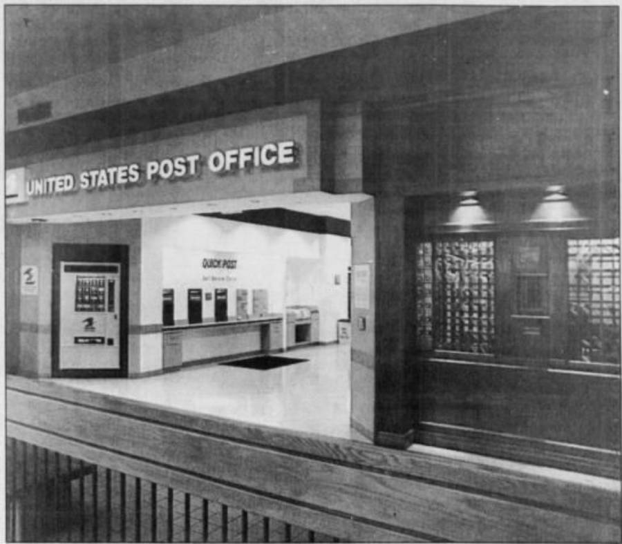 Black and white photo of a United States Post Office interior with mailboxes, service counters, and a sign above the entrance. The area appears empty and well-lit.