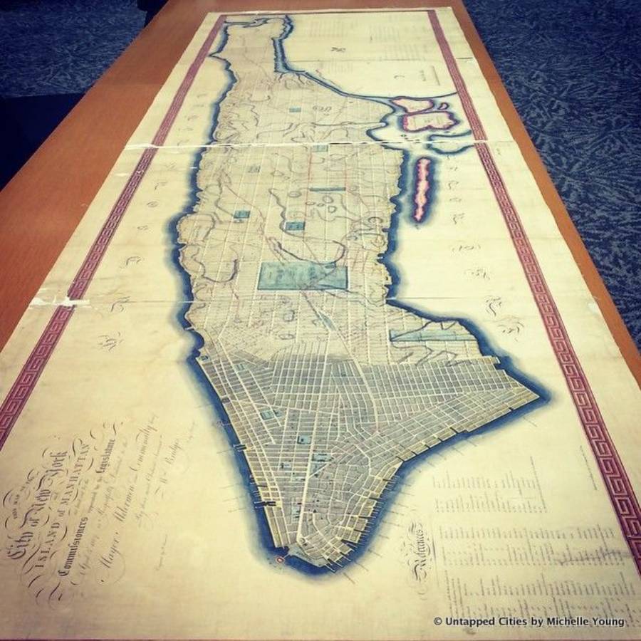 An old, detailed map of Manhattan is laid flat on a table, showing grid plans, parks, and land plots, surrounded by a decorative red border, with handwritten notes visible along one edge.