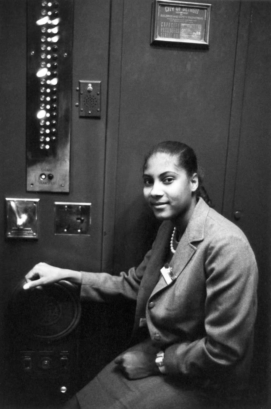 A woman in a suit sits in an old-fashioned elevator, hand on the control wheel, beside a panel with many buttons and switches, looking at the camera and smiling slightly.