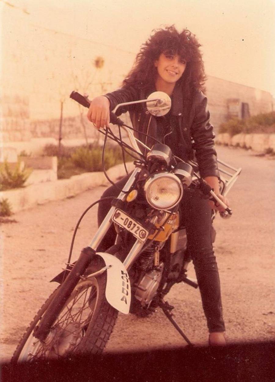 A person with curly hair and a leather jacket sits on a Honda motorcycle, smiling at the camera on a deserted, sunlit street with a stone wall in the background.