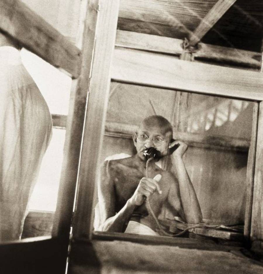 An elderly man with glasses sits behind a window, wearing minimal clothing, and speaking into a telephone. The setting appears modest and simple, with wooden beams and natural light coming through.