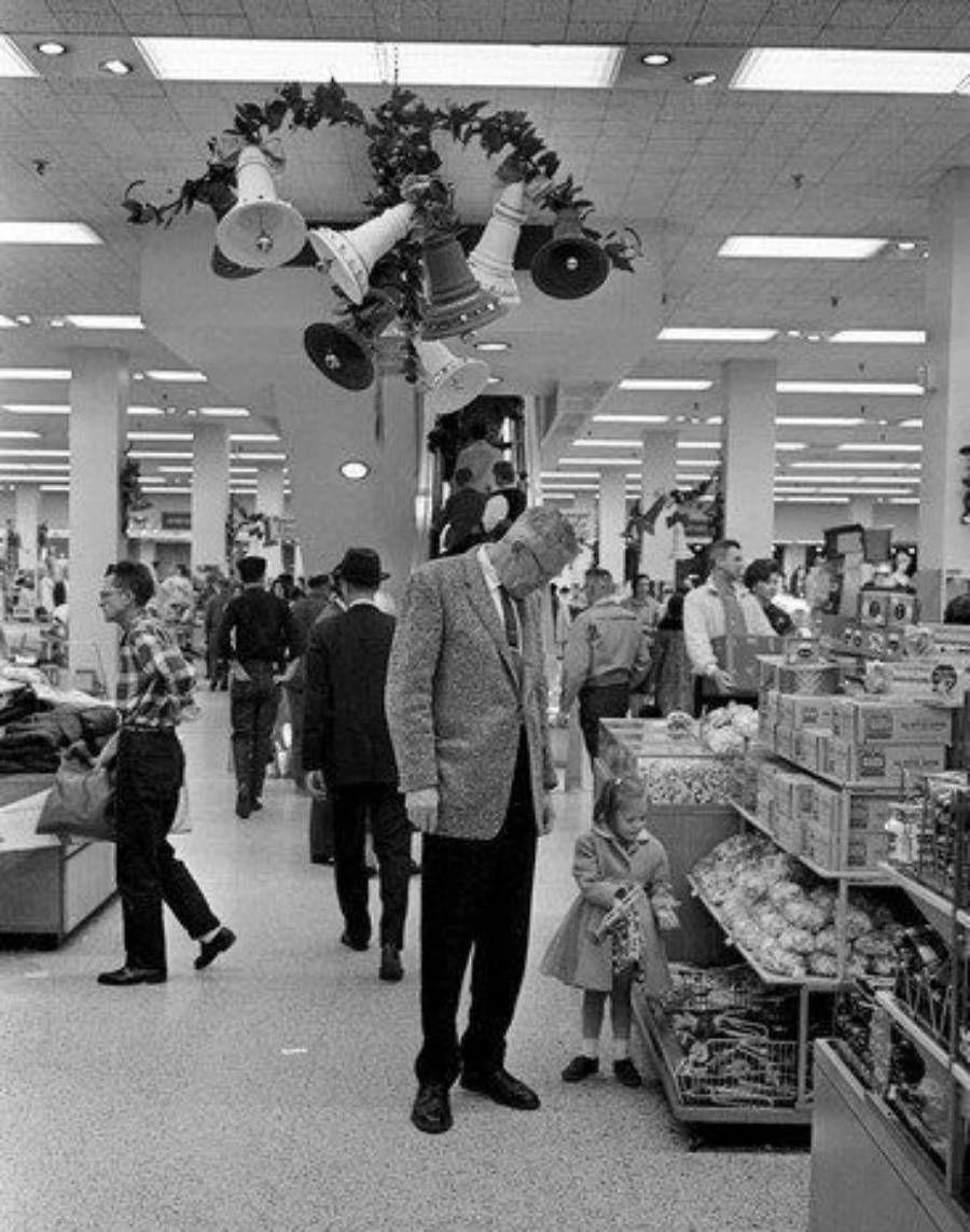 A man and a young girl look at toys in a busy department store decorated with garland and bells. Shoppers walk nearby, and shelves are filled with various products. The photo is in black and white.