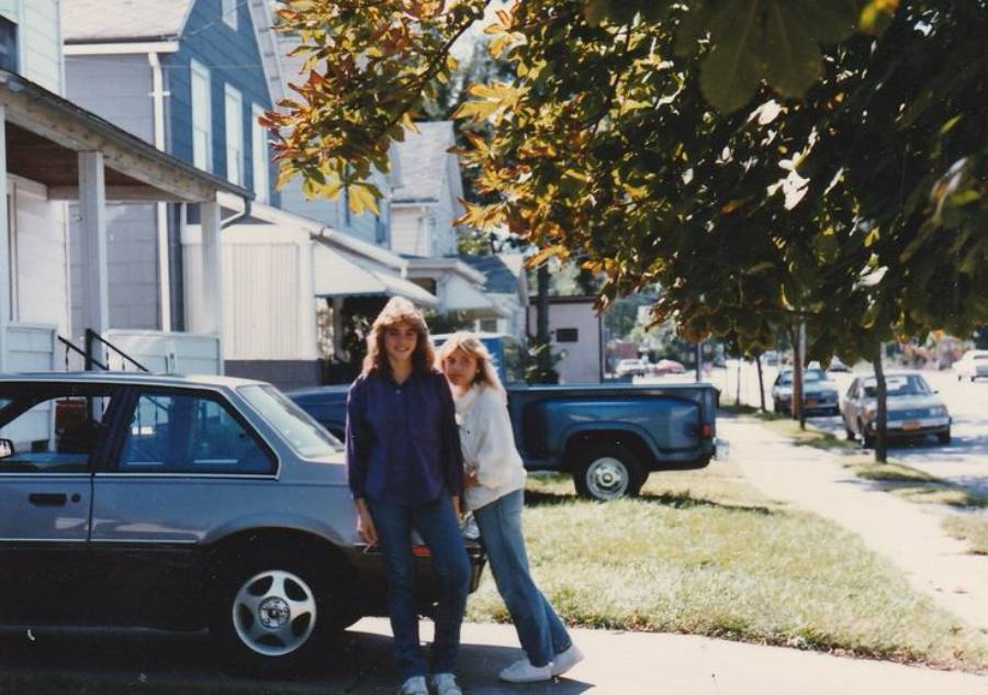Two people smile while standing on a sidewalk next to a parked car. Houses, trees, and more parked vehicles line the residential street in the background on a sunny day.