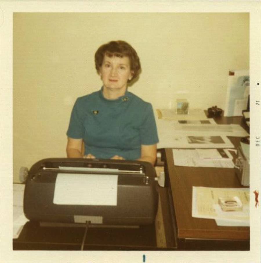A woman with short brown hair in a teal dress sits at a desk with a typewriter and paperwork, against a plain light-colored wall. The photo appears vintage, dated December 1971.